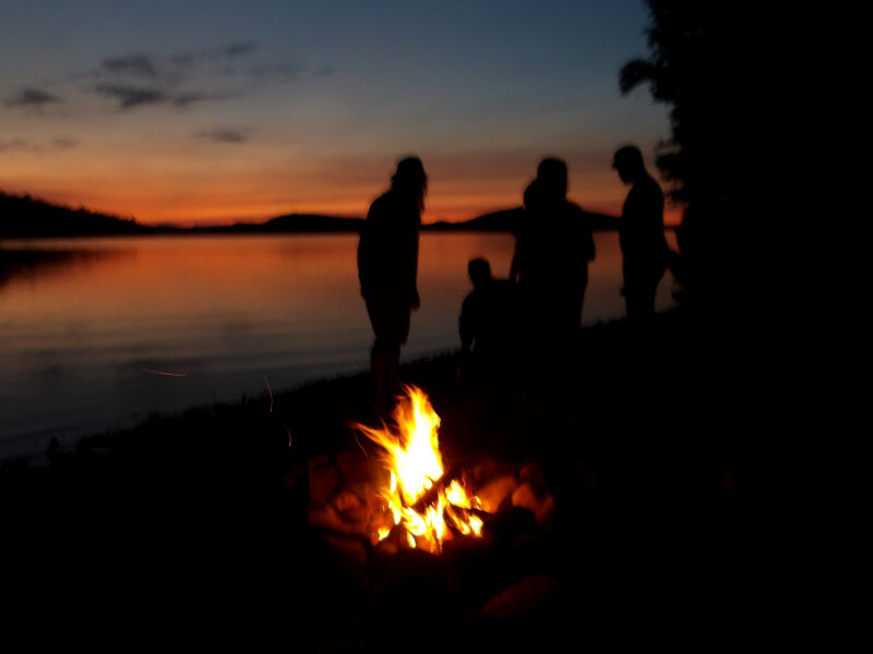 The image captures a serene evening scene. A campfire glows brightly in the foreground, casting light on silhouetted figures gathered nearby. The backdrop features a calm lake reflecting the vibrant colors of a sunset. The sky is painted with hues of orange and purple, creating a peaceful and inviting atmosphere.